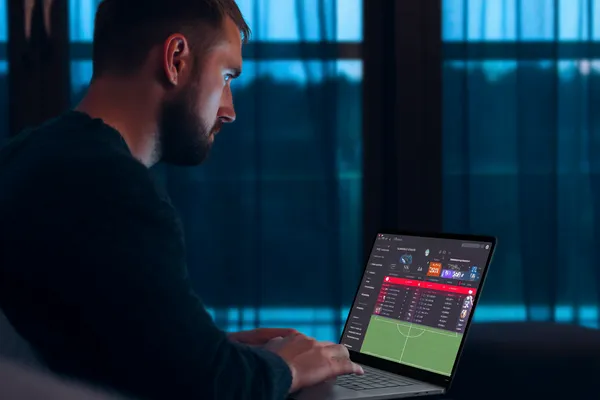 একজন পুরুষ তার ল্যাপটপে মনোযোগ দিয়ে কাজ করছেন, ব্যাকগ্রাউন্ডে লাইভ স্ট্যাটস — যা GOOD11111 মোবাইল অ্যাপের স্পোর্টস ও গেমিং ফিচারগুলোকে তুলে ধরে।